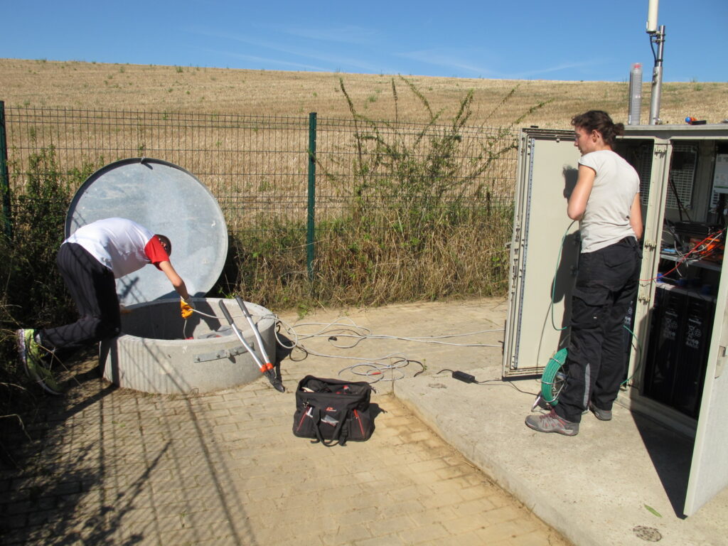 Maintenance des stations sismologiques : passage du câble dans le tuyau sous-terrain reliant la buse de puits à l’armoire grâce à un tire-fil (en vert)